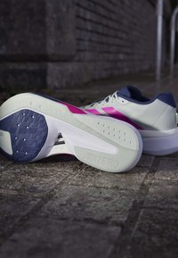 Paire de chaussures de course Adidas Adizero blanches et violettes posées sur un pavé en pierre, avec une chaussure montrant la semelle et le logo.