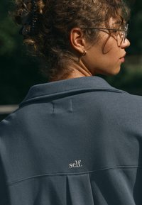 Sudadera gris oscuro con cuello, que presenta un sutil plisado en la parte posterior y el logotipo bordado "self." en un color claro. Textura suave.
