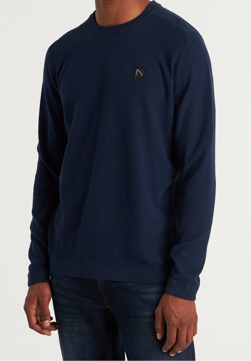 Pull à manches longues bleu marine avec un motif en maille texturée et un col rond côtelé. Présente un petit logo noir sur la poitrine.
