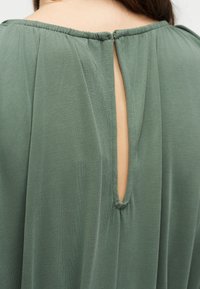 Κοντινή λήψη στο πίσω μέρος ενός πράσινου ενδύματος με μια μικρή οπή κλειδιού που ασφαλίζεται με κουμπί καλυμμένο με ύφασμα.