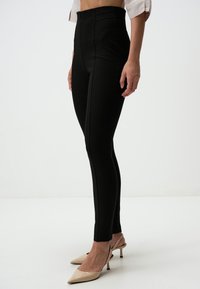 Leggings neri a vita alta con una texture morbida e una vestibilità sartoriale, caratterizzati da cuciture verticali e una silhouette pulita. Indossati con scarpe con tacco a punta.