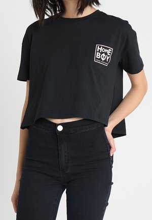 Nainen, joka pukeutuu mustaan lyhennettyyn T-paitaan, jossa on "HOME BOY" -logo, ja korkeavyötäröisiin mustiin farkkuihin, käsi lepää vyötäröllä.