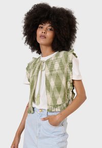 Gilet verde e crema con motivo a fantasia e bordi arricciati, legato davanti con frange, indossato sopra una maglietta bianca e una gonna di jeans azzurri chiari.