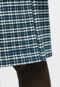 Ύφασμα με μπλε και λευκό καρό σε ρούχο, με μικρό κεντημένο λογότυπο πάνω από το γόνατο ατόμου με σκουρόχρωμο δέρμα.