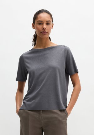 Kobieta o kręconych włosach, ubrana w szary top z krótkim rękawem i brązowe spodnie, stoi z rękami w kieszeniach na jednolitym tle.