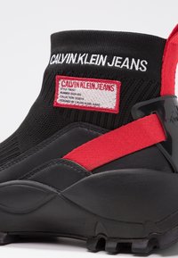 Černá vysoká teniska s červeným elastickým popruhem, logo Calvin Klein Jeans na pleteném kotníkovém límci a červený štítek s podrobnostmi o produktu.