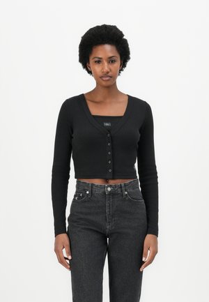 Cárdigan corto negro con mangas largas, cierre de botones, textura acanalada, usado sobre una camiseta negra de tirantes y combinado con unos vaqueros oscuros de tiro alto.