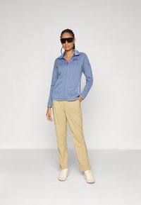 Giacca in pile blu chiaro con zip e finitura texturizzata, abbinata a pantaloni beige e scarpe da ginnastica di colore chiaro. Occhiali da sole neri come accessorio.
