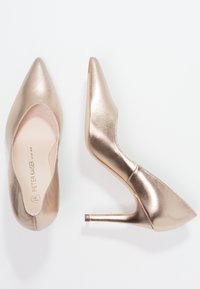 Une paire de chaussures à talons aiguilles métalliques dorées, l'une vue de dessus, l'autre vue de côté, sur un fond blanc.