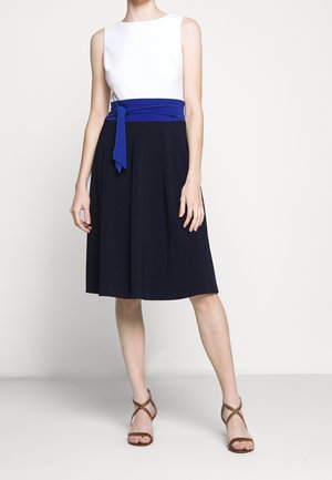 Biały top bez rękawów z niebieskim paskiem z materiału, w zestawieniu z granatową spódnicą do kolan. Gładka tekstura z lekko opadającym krojem. Brązowe sandały na paski.