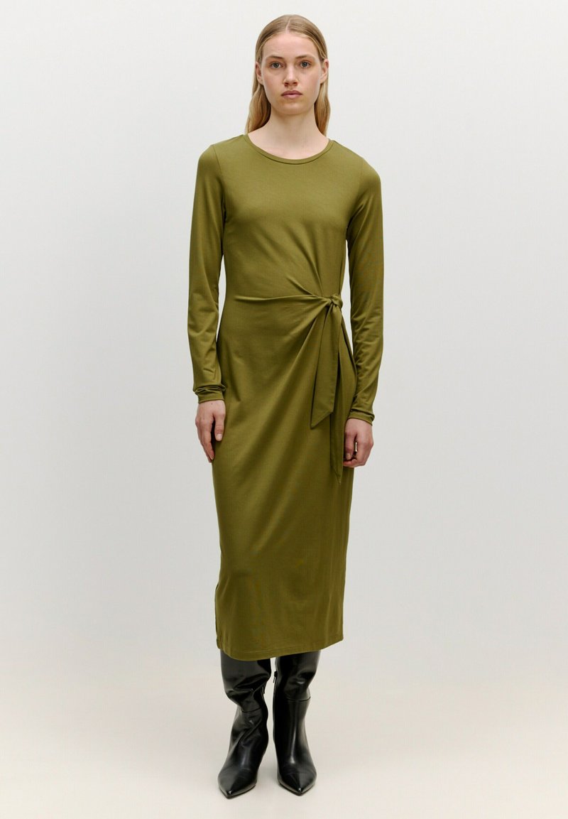 Olivgrünes, langärmliges Kleid mit rundem Ausschnitt und seitlichem Knoten-Detail, aus einem glatten Stoff gefertigt. Getragen mit kniehohen schwarzen Stiefeln.