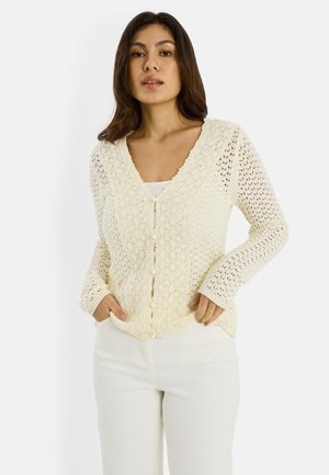 Mujer joven con un cárdigan de crochet color crema con botones, sobre una camiseta blanca, combinada con pantalones blancos, de pie con las manos en los bolsillos.