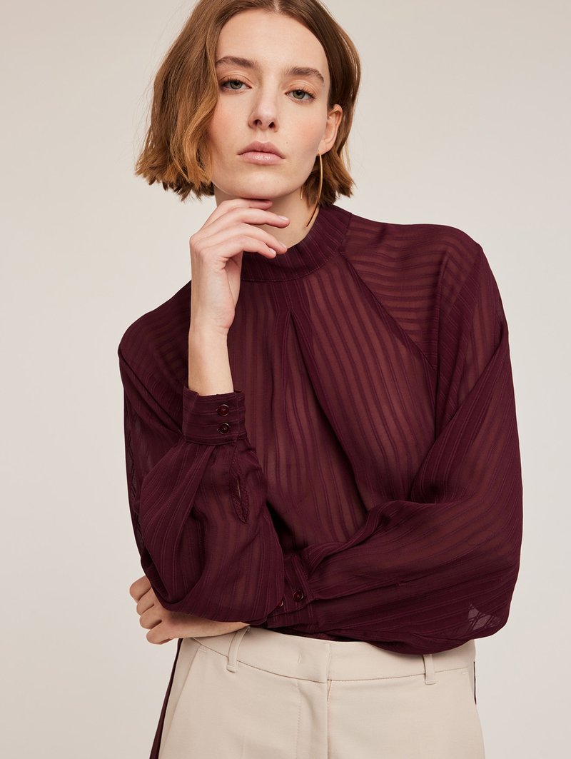 Diep bordeauxblauwe blouse met verticale strepen, hoge kraag en knoopmanchetten. Doorzichtig materiaal met een vloeiend ontwerp, gecombineerd met lichte broeken.