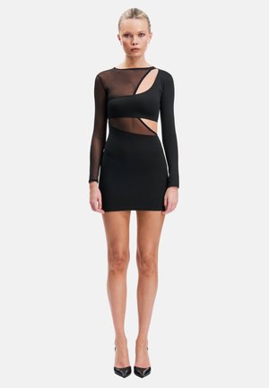 Femme debout portant une courte robe noire avec des panneaux en mesh transparent, des manches longues, des découpes et des chaussures à talons hauts noires sur fond blanc.