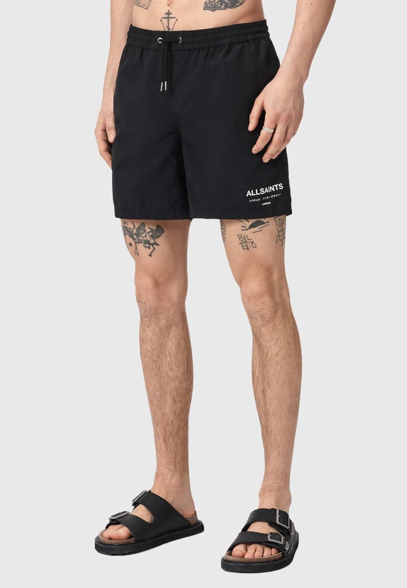 Man draagt zwarte AllSaints zwemshorts met trekkoord en zwarte sandalen, zichtbare tatoeages op benen en onderlichaam tegen een effen achtergrond.