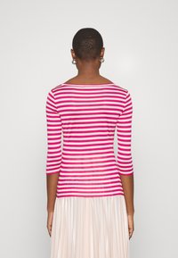 Person mit kurzen Haaren, die silberne Creolen, ein pink-weiß gestreiftes Langarmoberteil und einen hellen, plissierten Rock trägt und mit dem Rücken zur Kamera steht.
