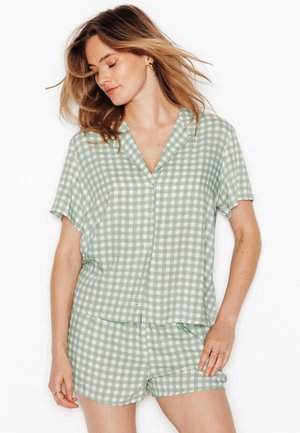 Femme aux cheveux mi-longs portant une chemise à manches courtes boutonnée à carreaux vert clair et blanc et un short assorti, debout devant un fond blanc.