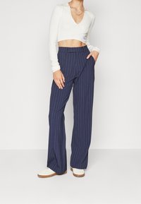 Biały, żebrowany crop top z dekoltem w serek i długimi rękawami, zestawiony z granatowymi spodniami w prążki o szerokich nogawkach. Sneakersy z białą cholewką i brązową podeszwą.