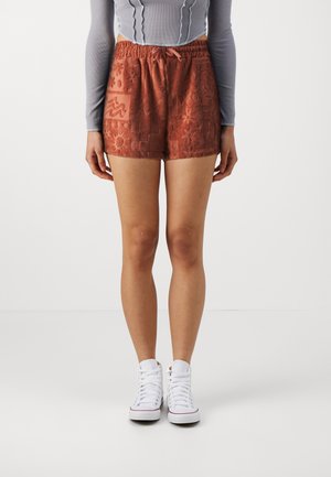 Frau trägt rostfarbene strukturierte Shorts mit elastischem Bund und Kordelzug, kombiniert mit weißen High-Top-Sneakers und einem langärmligen Crop-Top.
