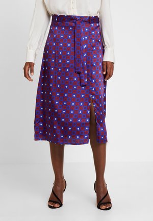 Femme portant une jupe midi à motifs bleus et rouges avec une fente à l'avant et une ceinture à nouer, associée à des talons ouverts noirs et un chemisier blanc.