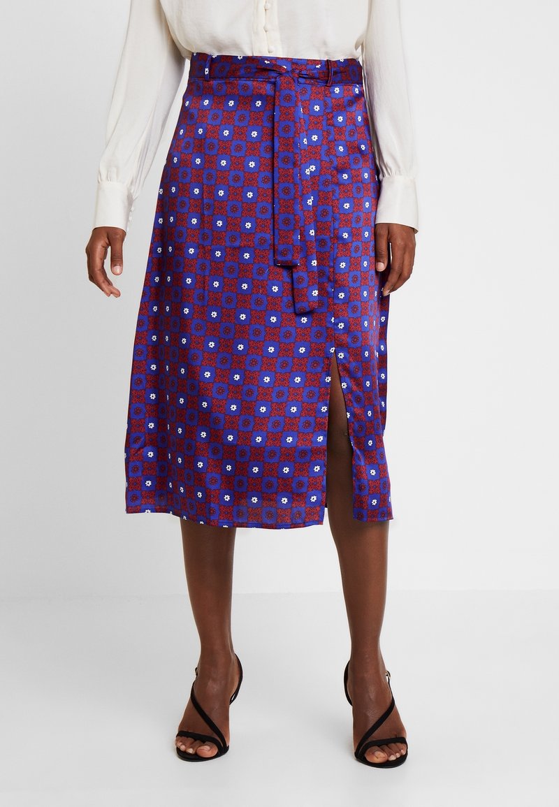 Femme portant une jupe midi à motifs bleus et rouges avec une fente à l'avant et une ceinture à nouer, associée à des talons ouverts noirs et un chemisier blanc.