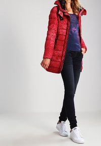 Mujer vestida con un abrigo acolchado rojo con patrón, camisa floral azul, jeans ajustados oscuros y zapatillas blancas, de pie con una mano en el bolsillo.