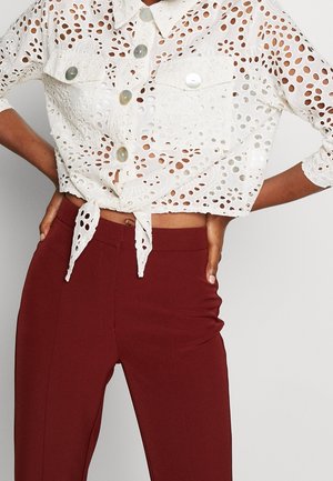 Biała koszula z haftem typu eyelet zapinana na guziki, związana w talii, zestawiona z wysokim stanem i dopasowanymi bordowymi spodniami, ręce spoczywają na biodrach.