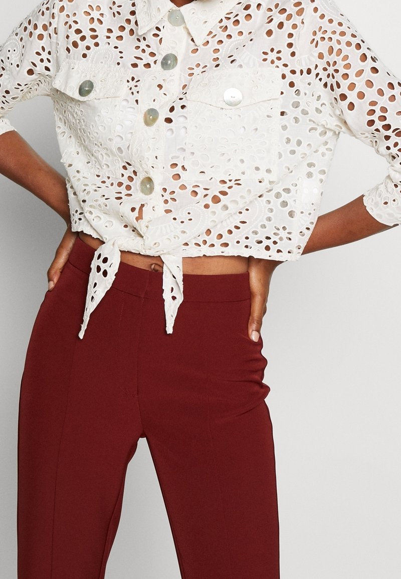 Chemise blanche en dentelle ajourée à boutons, nouée à la taille, associée à un pantalon ajusté taille haute couleur bordeaux, mains posées sur les hanches.