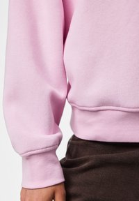 Gros plan d'une personne portant un sweat-shirt rose clair avec des poignets côtelés et un pantalon marron foncé.