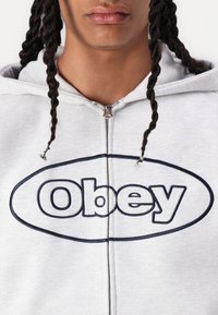 Sivý zipsový mikina s výraznou námornícko modrou značkou "Obey" na hrudi, má štandardnú kapucňu a textúrovanú látku.