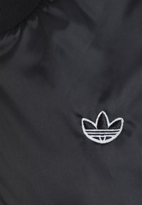 Svart tyg med en framträdande vit broderad logotyp med tre bladformer och tre horisontella ränder i basen. Slät textur.