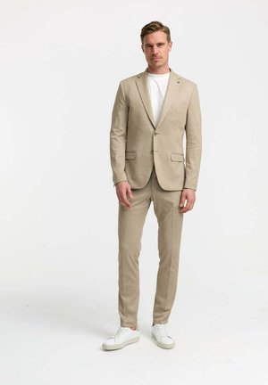 Beige Anzug aus strukturiertem Stoff, taillierte Blazerjacke mit zwei Knöpfen, passende schmale Hose, getragen mit einem weißen T-Shirt und weißen Sneakers.