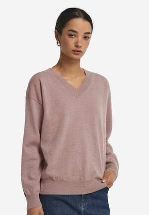 Light pink V-neck sweater made of soft fabric, featuring a relaxed fit, ribbed cuffs and hem, with a smooth texture. Worn over jeans.