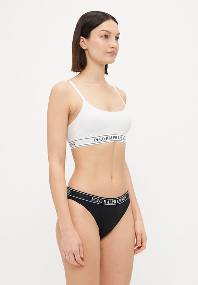 Hvit bralette og svart tanga undertøysett, laget av mykt stoff. Bralette har en logo-stripe; tangagen har en matchende logo-linning.