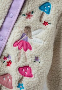 Fleece jaka ar purpursarkanām pogām, ar krāsainu izšuvumu, kurā attēlota pasaka, sēnes un ziedi uz krēmkrāsas fona.