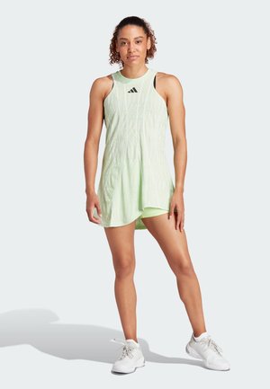 Robe de sport vert clair à design sans manches, avec un motif texturé et un ourlet arrondi. Des accents noirs et une paire de baskets blanches complètent la tenue.