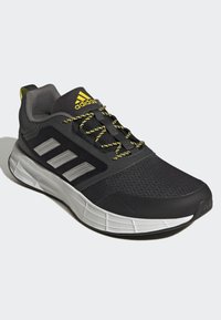 Zapatilla deportiva negra con acentos amarillos, materiales de malla y sintéticos, tres franjas plateadas, superficie texturizada y suela acolchada.
