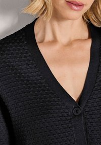 Close-up van een persoon die een gestructureerde zwarte cardigan met knopen en een V-hals draagt, waarbij een deel van de hals, het sleutelbeen en de lippen zichtbaar is.