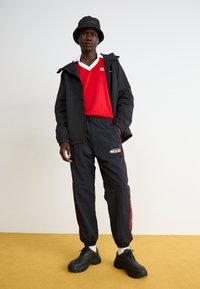 Sort vindjakke med hætte, rød v-hals skjorte, sorte bukser med røde detaljer, og chunky sorte sneakers. Glat tekstur, afslappet sportstil.