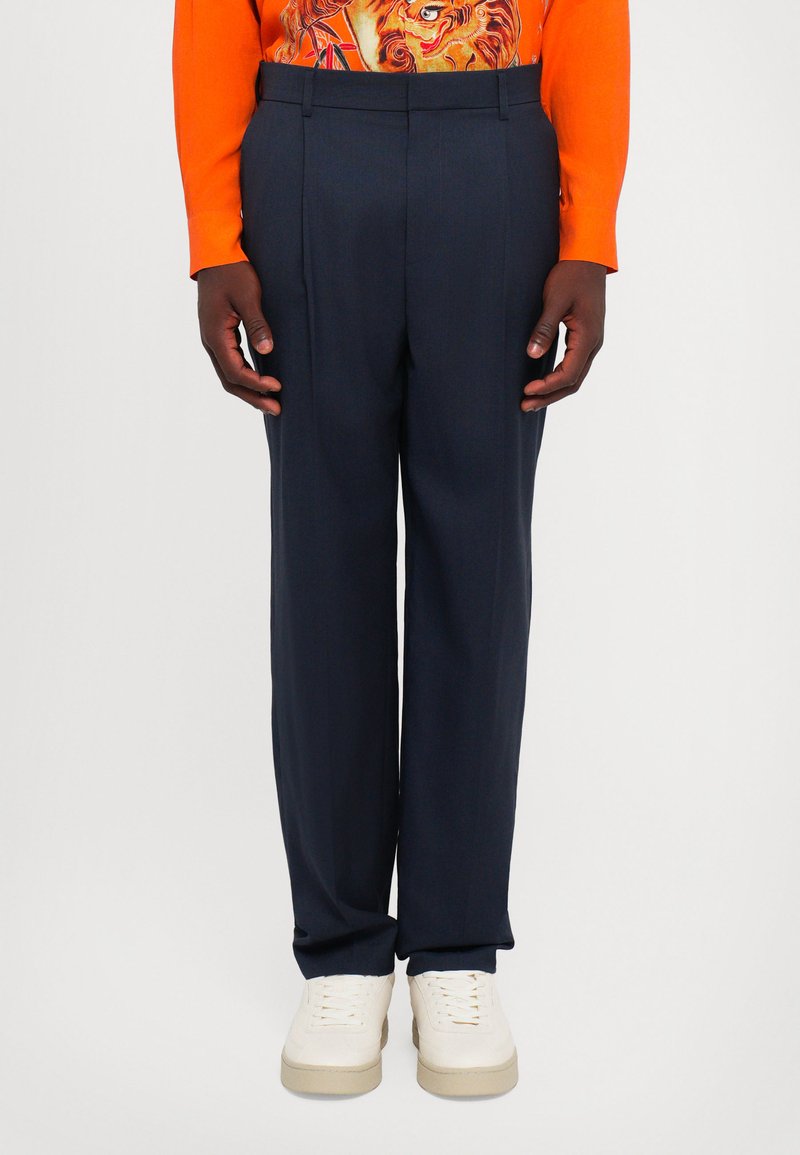 Mand iført navy skræddersyede bukser, en lys orange langærmet skjorte med grafik og hvide sneakers, stående på hvid baggrund.