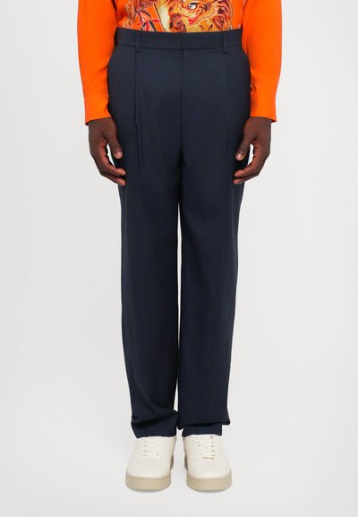 Marineblå bukser med skræddersyet pasform, med en glat tekstur og sidelommer, parret med cremefarvede sneakers. Lys orange langærmet top.