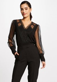 Blouse noire avec des manches transparentes à pois, des accents en dentelle et un décolleté en V avec un bord festonné. Assortie à un pantalon noir ajusté.