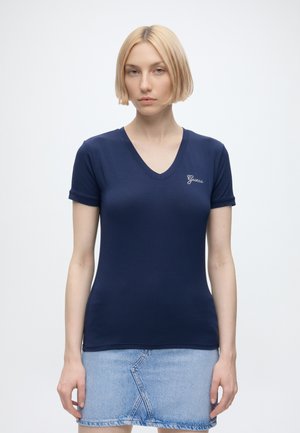 Giovane donna con capelli corti e biondi che indossa una T-shirt blu navy con scollo a V e una gonna di jeans azzurro chiaro, in piedi davanti a uno sfondo semplice.