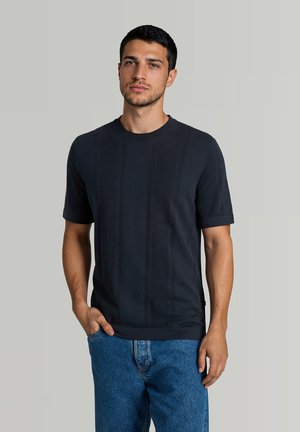 Ung mand med kort mørkt hår iført en sort kortærmet skjorte og blå jeans, stående med den ene hånd i lommen mod en ensfarvet baggrund.