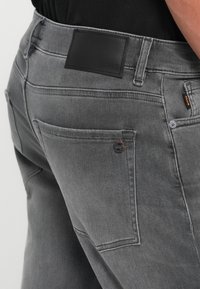 Šedé džíny z denimu s hladkou textúrou, s čiernou koženou nášivkou značky na páse a bočnou vreckom s oranžovým akcentom.
