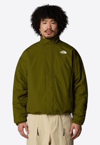 Uomo con capelli ricci indossa una giacca North Face verde oliva e pantaloni beige, in piedi davanti a uno sfondo bianco uniforme.