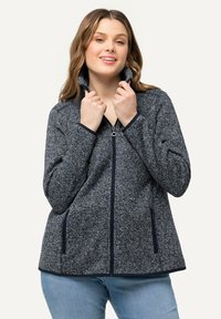 Grå fleecejacka med hög krage, dragkedjestängning och sidofickor. Kontrastfärgad marinblå kant längs kanterna och dragkedjan. Mjuk, texturerad tyg.