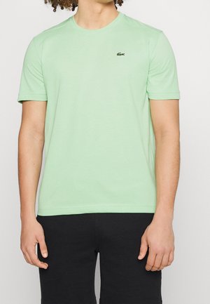 Osoba ubrana w jasnobrzeżowy T-shirt z krótkim rękawem z małym zielonym logo krokodyla na lewej piersi oraz czarne spodnie, stojąca prosto.