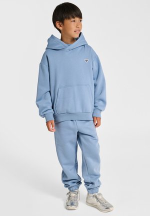 Jongen met een lichtblauwe hoodie en bijpassende joggingbroek met zilveren sneakers, staand en opzij kijkend tegen een witte achtergrond.