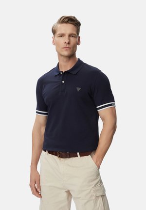 Uomo che indossa una polo blu navy con maniche a righe bianche e pantaloni cargo beige, in piedi con una mano in tasca.